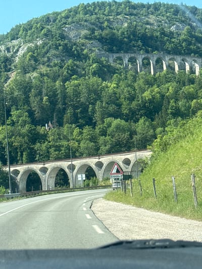 This is a photograph of the Oigney Viaduct in the city of Hauts de Bienne, France. The viaduct is made of stone arches and spans across a green, forested mountainside. In the foreground, a road curves to the right, with a grassy embankment on the right side of the road. There is a traffic sign with a mountain and "1000m" on it. In the background, the mountainside is covered in trees. The sky is clear and blue.