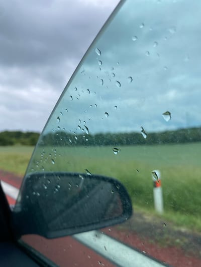 The image, taken from inside a vehicle, offers a view through a rain-streaked car window. The foreground shows the edge of the car's side mirror and the red and white stripe of the car door frame. The window itself is covered in numerous water droplets, distorting the view of the outside. Beyond the window, a blurred landscape unfolds, characterized by a green field or pasture, a line of trees in the mid-ground, and a cloudy, overcast sky. A white post with an orange band is visible in the distance, suggesting a roadside or open area. The overall impression is of a cool, possibly damp day, with limited visibility due to the rain. The location is stated to be Mirošovice, Czechia. There are no people or animals visible in the image.