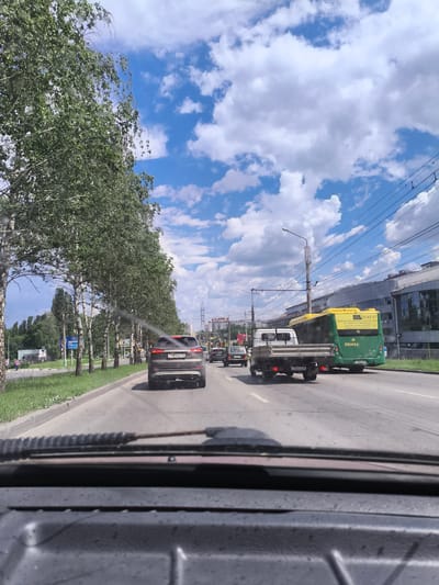 The image captures a view from inside a vehicle looking out at a street scene in Malaya Valyaevka, Russia. A line of birch trees with lush green foliage borders the right side of the road, which is paved with asphalt and marked with white lane lines. In the foreground, the dashboard and windshield of the vehicle are visible, with what appears to be water droplets or smudges on the glass.
Several vehicles are driving on the road. A dark-colored SUV is in the right lane, followed by a flatbed truck. Further ahead, a green and yellow bus is also in motion. The bus has writing on its back, including "ПЕНЗА" (Penza) and a date. In the distance, buildings and power lines can be seen. The sky is a vibrant blue, dotted with fluffy white clouds. A pedestrian is visible on the left side of the road, walking near a sign. The overall impression is a typical daytime scene on a suburban or urban street in Russia.