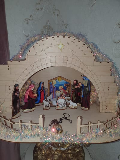 A nativity scene is displayed against a textured wall with ornate wallpaper. The nativity scene is constructed from light-colored wood and features several figures depicting the birth of Jesus. In the center, a manger scene is visible with Mary, Joseph, and the infant Jesus. To the left, figures in traditional attire, including one kneeling and presenting a gift, stand near two sheep. To the right, two figures are also present, one appearing to offer a gift. Above the figures, an arched structure made of wooden blocks creates a stable-like appearance, adorned with tinsel and small star cutouts. A string of delicate lights is woven through the structure. The foreground of the scene includes a small wooden fence, also decorated with tinsel. The overall setting is domestic.
