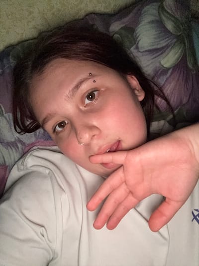 An eye-level, close-up portrait features a young person lying down indoors, head tilted. The subject gazes directly at the viewer with dark brown eyes. Their right hand partially covers their mouth and nose, with the tongue subtly extended between the fingers, creating a playful or shy expression.

The subject has dark hair and several piercings: two small barbell piercings are visible above the left eyebrow (from the viewer's perspective), and a simple hoop piercing adorns the left nostril. They are wearing a light-colored, possibly off-white, long-sleeved top. A faint, dark blue graphic, appearing as a stylized 'X' or intersecting lines, is visible on the upper right side of the garment.

The background shows bedding with a dark, intricate pattern, incorporating shades of purple, green, and blue, possibly depicting floral or abstract designs. The lighting suggests an indoor setting, likely soft and ambient, without indicating a specific time of day. The image primarily captures a casual and intimate moment. No explicit location identifiers are visible within the image, beyond an indoor environment. The provided context states the location is Lyman, Ukraine.