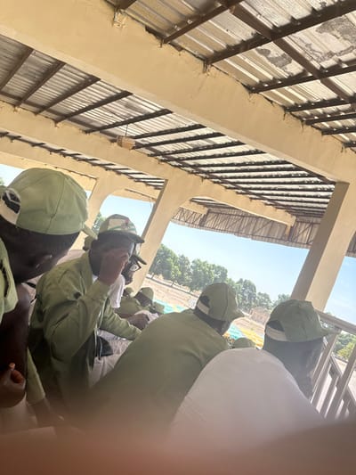 The image shows approximately six individuals gathered under a rudimentary shelter in Ajiwa, Nigeria, viewed from slightly above and behind. The shelter features a corrugated metal roof supported by light-colored concrete beams and pillars. Most individuals are wearing light olive-green long-sleeved uniforms and matching caps, consistent with the Nigerian Youth Service Corps (NYSC) attire. One individual in the mid-ground, wearing glasses and a cap with a visible emblem (likely the Nigerian flag and eagle), is seen touching their face or forehead. The background reveals an open, outdoor area under a clear blue sky, with sparse green trees and what appears to be terraced or stepped ground in the distance. A white, out-of-focus object occupies the lower foreground, possibly part of the camera operator's clothing. A small, square, light-colored fixture hangs from one of the overhead beams.