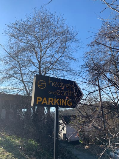 The image, rotated clockwise, captures an outdoor scene dominated by a black sign partially framed by bare tree branches against a clear blue sky. The sign is rectangular with a pointed top edge and displays text in white and gold. Vertically arranged on the left side of the sign are the words "heaven and Earth," with "heaven" in a smaller, stylized font above "and Earth." A circular logo, resembling a stylized "H," is positioned to the left of these words. To the right, in large, vertically oriented golden letters, the word "PARKING" is prominently displayed.
The sky is bright and a vibrant blue, indicating sunny weather during the day. Numerous bare deciduous tree branches, devoid of leaves, extend across the frame, suggesting a late autumn, winter, or early spring season. In the background, behind the sign and trees, glimpses of a building or structure are visible, along with some sparse vegetation and what appears to be a paved area or road. No people or specific activities are depicted, portraying a serene, static landscape in Kastraki, Greece.