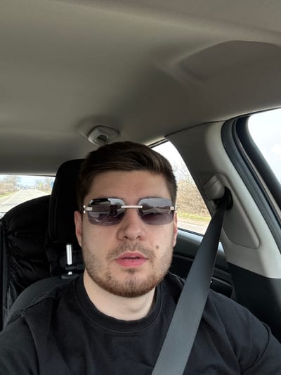 A male individual is depicted in a selfie taken from the interior of a vehicle, asserted to be in the city of Novi Chiflik, Bulgaria. The subject, of light complexion, has short dark brown hair and a light brown beard, and is wearing a plain black crew-neck t-shirt. He is wearing rimless rectangular sunglasses with dark lenses and silver-colored metal components, including visible "Cartier" branding on the left temple. His mouth is slightly ajar. He is seated, secured by a black vehicle seatbelt crossing his chest and left shoulder. The vehicle interior shows a light grey headliner and a dark seat. Visible through the vehicle's windows are an outdoor environment featuring a paved road extending into the distance, lined with bare trees and shrubs under an overcast sky. A vehicle's "AIRBAG" designation is visible on the right-side interior pillar.