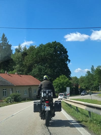 The image shows a rear view of a motorcyclist on a paved road in Strunkovice nad Volyňkou, Czechia. The motorcycle is equipped with two large black panniers on either side and a top box. The license plate reads "6P 5224". The motorcyclist is wearing a black jacket and a yellow and black helmet. To the left of the road, there is a light-colored building with a red tiled roof and a grey roof in the foreground. A wooden fence and grass are visible to the right of the road, followed by a metal guardrail. Trees and a clear blue sky with a few white clouds form the background. In the distance, another vehicle, a white car, is visible on the road, as well as a silver SUV. There are also road signs visible, including a railway crossing sign. The scene appears to be a sunny day.