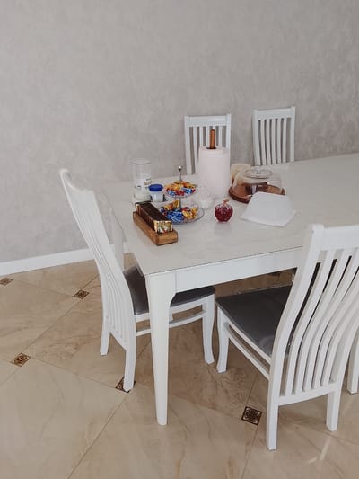 A sparsely populated indoor scene shows a white dining table with three chairs, two facing the viewer. On the table are various items, including a tiered serving tray of candies, a wooden tray filled with small packets, a roll of paper towels, and a covered cake stand. An apple and a white disposable food container also sit on the table. The table and chairs are set on a light-colored tile floor with dark accent tiles. The location is Assinovskaya, Russia.