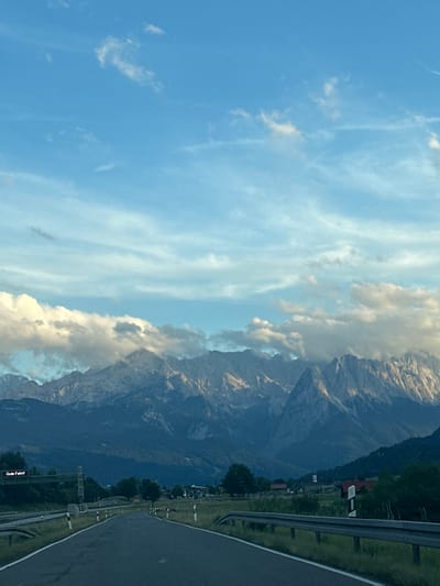 The image depicts a scenic view of the Alps in the vicinity of Farchant, Germany. A paved road curves towards a majestic mountain range partially obscured by clouds. The sky is a clear blue, with wispy clouds scattered across it. The mountains are rugged and imposing, with their peaks catching the light.
In the foreground, the road is bordered by guardrails and grassy verges. To the left of the road, an overhead sign displays the German phrase "Gute Fahrt!", which translates to "Have a good trip!". The environment appears to be a rural or semi-rural area, with some scattered buildings visible in the distance. The overall impression is one of a serene and picturesque landscape, ideal for a road trip.