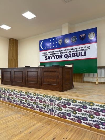 The image depicts an indoor scene, likely a room or hall in Zafarabad, Uzbekistan, with a ceremonial or formal atmosphere. Dominating the central area is a dark wooden podium or cabinet with multiple paneled doors, suggesting it's a piece of furniture for a speaker or presenter.

Behind the podium, a large banner is displayed. The banner has a blue section on the left, adorned with various official emblems and stars, and a crescent moon. To the right of the blue section is a white stripe with prominent text in a dark font. To the right of the white stripe is a vibrant green section. The text on the banner reads: "JIZZAKH VILOYATI MUHOFAZA QILUVCHI VA NAZORAT ORGANLARI RAHBARLARINING OMMAVIY SAYYOR QABULI". This translates to "Public Reception of the Heads of the Protection and Oversight Bodies of Jizzakh Region". This text clearly indicates that the event is a public reception hosted by the protection and oversight bodies of the Jizzakh region.

To the right of the podium and banner, a decorative textile, possibly a traditional Uzbek ikat fabric, runs diagonally across the frame. This fabric features a repetitive pattern of stylized floral or eye-shaped motifs in various colors, including purple, green, yellow, and dark blue, on a white background. It is laid out on a wooden surface, which appears to be part of the room's architecture or flooring, made of light-colored wooden planks arranged diagonally.

The ceiling is visible at the top of the image and features modern square light fixtures, one of which is illuminated, casting a bright white light. The walls appear to be painted a light, neutral color. The overall lighting suggests it is daytime, and the indoor setting indicates controlled environmental conditions. The scene conveys a sense of official proceedings and cultural presentation.