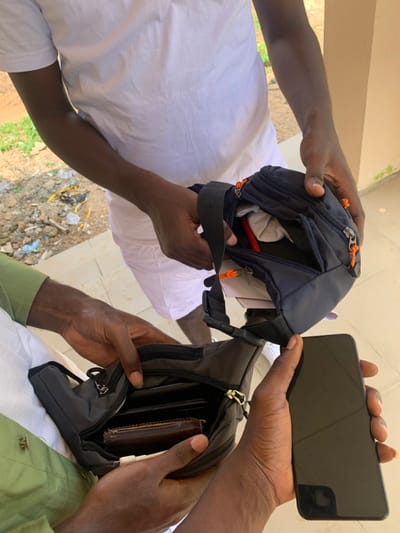 The image captures three individuals in an outdoor setting in Kubi A, Nigeria. The central figure, wearing a white t-shirt and white trousers, holds a black fanny pack with orange zipper pulls open, revealing a crumpled white item and a red cylindrical object within. A second individual, on the lower left and wearing a green shirt with white trousers, holds a similar black fanny pack with silver zipper pulls open, displaying a brown leather wallet and other dark objects inside. A third individual, on the lower right, holds a dark-colored smartphone with a blank, reflective screen, positioned towards the fanny packs. The ground is paved, and the background shows a dirt area with scattered refuse and some vegetation to the left, and a light-colored building wall with green discoloration to the right.