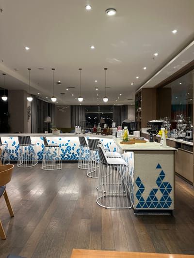 The image displays the interior of an unoccupied bar or lounge area, located in Zeytinburnu, Turkey. A prominent, curved bar counter occupies the central and left portions of the frame. Its facade is white with a repeating pattern of blue triangles. Along the bar are multiple high-backed bar stools with white wireframes and grey seat cushions.

To the right, a service counter is equipped with an espresso machine, a blender, various glassware, and a yellow spray bottle. On the bar surface, objects include planters with greenery, several decorative bottles, and a pitcher of lemon-infused water on a wooden tray. The flooring is dark wood, and the white ceiling contains numerous recessed light fixtures and several spherical pendant lamps. Large windows in the background overlook a dark urban exterior with distant lights. No individuals are present.
