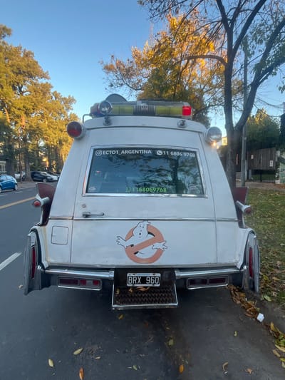 A white modified Cadillac hearse, resembling the "Ecto-1" vehicle from the Ghostbusters movie, is parked on the side of a road. The vehicle has the iconic Ghostbusters logo on the rear passenger side. Its license plate reads "BRX 960". A sticker on the back window shows "@ECTO1_ARGENTINA" and the Argentinian phone number "+11 6806 9768." The location is likely in Martínez, Argentina, based on the license plate and the sticker. The surrounding environment includes trees and a glimpse of residential buildings. The sky is clear blue. Another vehicle is parked ahead on the street.