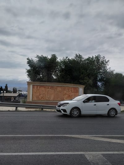 The image shows a white sedan driving on a paved road in Üşaral, Kazakhstan, under an overcast sky. To the left of the road is a low metal guardrail, and behind it, a large relief mural is visible. The mural depicts figures and designs in warm, earthy tones. Behind the mural, there are trees with dense foliage and some structures, including what appears to be a covered market or roadside stalls with lamp posts. In the far background, mountains are faintly visible. The car is the primary subject in the foreground, captured in profile as it moves along the road. A person is visible inside the car, driving.