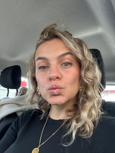 A young woman with light brown curly hair and green eyes is the main subject of the image. She has fair skin and is wearing earrings and a gold necklace with a large medallion. She is wearing a black top, possibly a sweater, and is seated in what appears to be the passenger seat of a vehicle. The interior of the vehicle is predominantly gray.
Through the windshield, a partially visible sign or poster with the word "Energies" can be seen. Based on the visual cues, the location is in Valence, France. The presence of a vehicle suggests the subject is either parked or driving. The expression on her face is a 'duck face'.