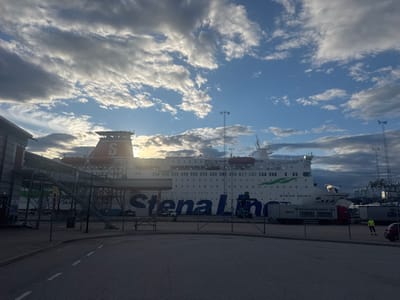 A large Stena Line ferry, named "Stena Spirit", is docked at a port. The ferry is white with a prominent red funnel bearing the Stena logo. Large blue lettering on the side of the ferry reads "Stena Line". The scene is set in a harbor with industrial elements such as cranes and shipping containers visible in the background. A red truck with the text "SPRENGNINGSMATTER" and "BERG44" on its side is parked near the ferry. A person wearing a yellow high-visibility vest stands on the tarmac. The sky is dramatic with a mix of bright sunlight breaking through dark, textured clouds, suggesting either sunrise or sunset. The overall atmosphere is one of activity at a port. The time of day appears to be either early morning or late afternoon. The location is likely Karlskrona, Sweden, given the provided context.