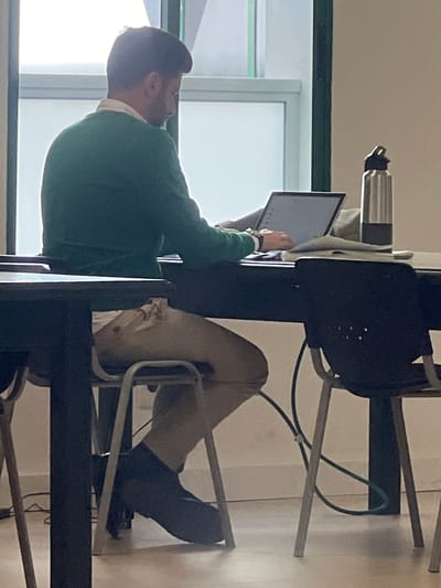 The image, taken in La Unión, Spain, depicts an indoor scene focused on a male individual seated at a dark rectangular table. The individual is seen from the upper back, facing a large, brightly lit window with white framing and a dark green vertical mullion. He wears a green long-sleeved sweater over a white collared shirt, light-colored trousers, and dark footwear. Both hands are positioned on an open silver laptop computer, indicating active use, possibly typing or operating the trackpad. To the right of the laptop, a silver insulated water bottle with a black lid rests on the table. Papers or a thin notebook are also visible on the table surface. The individual is seated on a chair with a dark seat and a light-colored metal frame; a second identical chair is positioned to the right. A dark cable is visible on the light-colored floor beneath the table. The walls are light-colored.