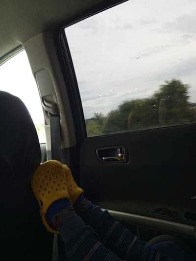 The image shows a child's feet, wearing yellow Crocs and striped socks, resting on the interior of a car. The child is likely a passenger, with their feet positioned in a relaxed manner. The scene appears to be a car interior looking out a window. Outside the window, there are trees and a cloudy sky, suggesting it is daytime. The overall mood is relaxed and possibly indicative of travel. There is no visible text in the image.