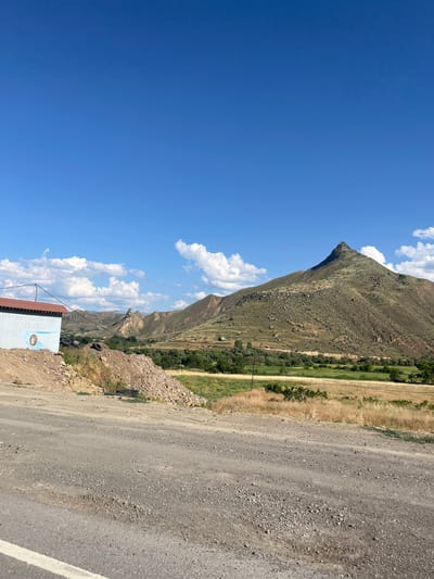 The image depicts a scenic mountain landscape in Gladzor, Armenia, under a clear blue sky dotted with fluffy white clouds. The scene is oriented vertically, with the sky occupying the left two-thirds of the frame and the mountainous terrain and a dirt road on the right.

The foreground features a rough, gravelly dirt road, likely a rural path, with a white line painted on its surface, suggesting it might be a seldom-used or older road. To the left of the road, a small, utilitarian building with a reddish-brown roof is visible. The side of this building is painted with light blue and white, featuring some text and a circular emblem. The text is partially obscured but appears to be in a non-Latin script.

Beyond the road and building, the landscape transitions into a steep, rocky mountainside covered with sparse vegetation. The mountain rises dramatically, showcasing its rugged texture and geological formations. Further into the distance, a valley unfolds, with patches of green fields and scattered trees, hinting at agricultural activity or a natural, verdant area within the mountainous region. The overall impression is one of a bright, sunny day, likely midday given the strong light and clear sky, in a remote and natural setting. There are no people visible in the image. The mood is peaceful and serene, highlighting the natural beauty of the Armenian countryside.