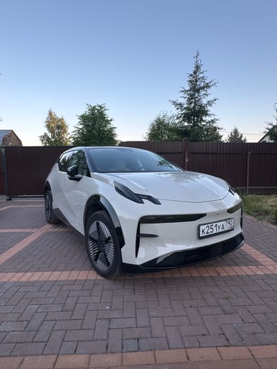 In Борисьево Днп, Russia, a white electric car, identified by the license plate K251YA 790, is parked on a brick-paved driveway. The car, a sleek, modern hatchback, is positioned at an angle facing the viewer, with its front end slightly turned towards the right. The driveway is bordered by a dark brown wooden fence, behind which several trees and a portion of a building are visible. The sky above is a clear, pale blue, indicating daytime, possibly late afternoon or early evening, given the soft lighting. The scene is a residential or semi-rural environment, with the car being the primary subject.