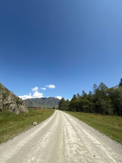 A bright, sunny day in Bichiktu-Boom, Russia. The image shows a dirt road stretching into the distance, flanked by green grassy fields. On the left side of the road, a rocky, moss-covered hillside rises. In the middle distance, a small, red-roofed building sits in a field. Behind the field, a range of majestic mountains are visible, their peaks partially obscured by fluffy white clouds against a brilliant blue sky. On the right side of the road, a dense forest of evergreen trees creates a dark, verdant border. The road itself is wide and appears to be made of light-colored gravel and dirt, with tire tracks visible. The scene conveys a sense of open space, natural beauty, and rural tranquility.