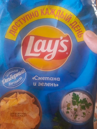 The image is a close-up of a blue bag of Lay's potato chips. The bag features the iconic Lay's logo in red and white on a yellow background. Above the logo, a yellow banner with red text reads "ДОСТУПНО КАЖДЫЙ ДЕНЬ," which translates to "AVAILABLE EVERY DAY" in Russian. Below the logo, in Russian, it says "«Сметана и зелень»," meaning "Sour cream and greens."
In the bottom left corner, there is an oval emblem with Russian text that reads "НАТУРАЛЬНЫЙ ОТБОРНЫЙ КАРТОФЕЛЬ," indicating "NATURAL SELECT POTATOES." A portion of the chips are visible in a dark bowl in the lower left, with a date stamp visible on the bag below them. In the lower right, there is a picture of a bowl of sour cream dip garnished with parsley. The overall impression is a product shot of a specific flavor of Lay's chips. The lighting suggests it's likely indoors, possibly a product photo shoot.