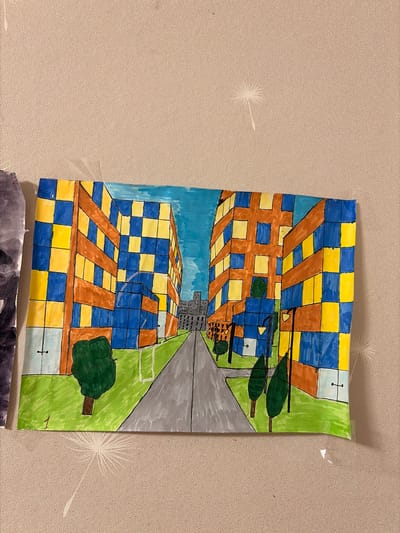 The image shows a child's drawing of a city street with buildings. The buildings are colorful, with alternating blue and yellow squares on their facades. There are trees and lampposts along the street, and a grey road leads into the distance. The sky is blue. The drawing is taped to a textured wall with a dandelion-like pattern. The drawing is located in Leninogorsk, Russia.