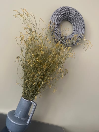 The image shows a close-up view of dried yellow flowers in a grey ceramic vase. The vase is tilted and rests on a dark surface. Behind the vase, a grey woven wreath is hanging on a light grey wall. The lighting suggests an indoor setting. No people or actions are depicted in the image. The image does not provide any information about the location of Староживотинное, Russia.