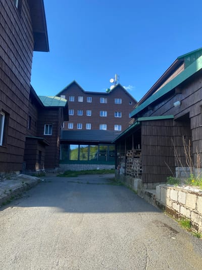 The image depicts a view of a wooden building complex in Loučná nad Desnou, Czechia, under a clear blue sky. The buildings, constructed with dark wood siding, feature multiple windows and green trim on the roofs. The tallest building in the center exhibits a gabled roof and a communication antenna mounted on top. A paved walkway runs alongside the buildings, with a stone retaining wall and sparse vegetation visible along the edges. Pallets stacked under one of the structures can be seen.