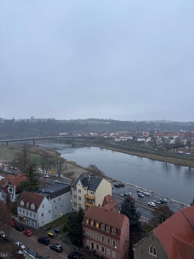The image captures an elevated, panoramic view of a town or city, likely Meissen, Germany, overlooking a river on a heavily overcast day. The perspective is from above, looking down towards the river and the landscape beyond. The sky is a uniform, pale grey, indicating a completely cloudy day with low light conditions, possibly late autumn or winter due to the bare trees.
A wide river, appearing calm and reflecting the grey sky, flows from the foreground towards the distant horizon, creating a prominent horizontal element across the middle of the scene. Along the near bank of the river, a paved road runs parallel to the water. Numerous cars are parked along this road, and some appear to be driving, though no individual people are clearly visible. A collection of buildings, primarily multi-story structures with varying architectural styles, some featuring red-tiled roofs and others darker, flat roofs, line this near bank. Warm lights are visible in some windows, suggesting either late afternoon or interior lighting due to the dim day.
On the far side of the river, another line of buildings, smaller and more interspersed with vegetation, follows the riverbank. Beyond these structures, a tree-covered hillside rises, showing patches of dull green and brown, extending into the misty distance where the landscape fades into the grey sky. A bridge with visible arches spans the river further downstream (towards the left of the image). The overall atmosphere is muted and somewhat somber, characterized by the grey tones of the sky, river, and damp-looking ground. No discernible text is visible.