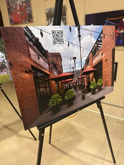 A photographic print on display shows a brick-clad streetscape with red awnings above outdoor dining. Planters filled with greenery line the sidewalk. Tall lampposts with hanging lights stretch down the street, under a partly cloudy sky. A QR code is affixed to the bottom left corner of the print. The print rests on a black easel.