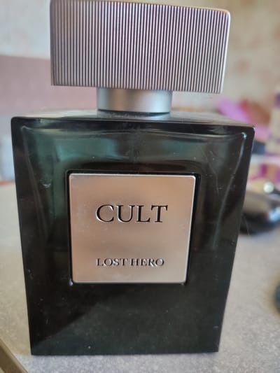 The image shows a close-up, eye-level, indoor shot of a black glass perfume bottle with a silver cap. The bottle has a silver rectangular label with raised text that reads "CULT" at the top and "LOST HERO" at the bottom. The background is blurred and appears to be an indoor setting with a light-colored wall and some out-of-focus objects. The image quality is fair, with some dust or smudges visible on the bottle and cap. The location of this image in Salavat, Russia, is not discernible from the visual content.