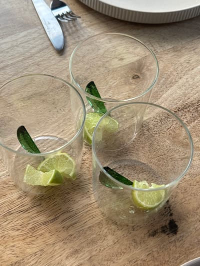 On a light-toned wooden surface in Culburra Beach, Australia, three clear glass tumblers are arranged. Each tumbler contains between one and three freshly cut green lime wedges and a single dark green, leaf-shaped stirring stick. Small droplets or residues are visible within the glasses. In the upper left quadrant, a silver dinner knife and fork are present, with the knife to the left of the fork. A portion of a round, off-white ceramic plate with a ribbed edge is visible in the upper right quadrant. The wooden surface exhibits a discernible grain pattern and minor dark discolored areas. No individuals are present.