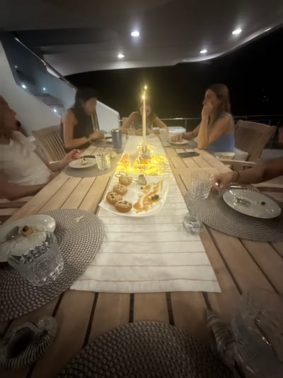 A group of people are gathered around a wooden table laden with food and drink, likely on a boat, given the hull and lights visible on the left side of the image. The setting appears to be a nighttime meal or gathering. The location is unknown, but could be Mexico.
The table is decorated with a striped runner, placemats, plates, and glasses. A dessert platter with small pastries and caramel drizzle sits in the center. The presence of candles suggests a celebratory or relaxed atmosphere.
Several individuals are visible around the table. One woman, wearing a blue top, is seen leaning over the table, possibly interacting with the food or other guests. Another woman in a black top and a person in a white dress are partially visible.
The scene captures a social gathering with food, drinks, and conversation, possibly on a boat at night.
