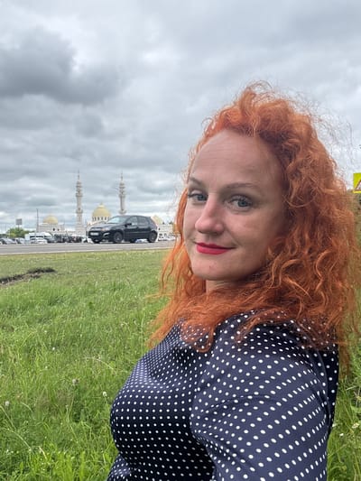 A woman with long, curly red hair and blue eyes smiles at the camera. She is wearing a navy blue polka dot dress. Her makeup includes red lipstick. The photo is taken from a slightly elevated angle, looking down at her.

In the background, there is a grassy field and a road with several cars. A black SUV is prominently parked on the side of the road. Further in the distance, several white minarets and golden domes of what appear to be mosques are visible against a cloudy sky. The overcast weather suggests it might be a cloudy day. The scene is set outdoors in a location that features both natural elements (grass, sky) and man-made structures (road, cars, religious buildings). The overall impression is of a casual outdoor setting, possibly during the daytime.