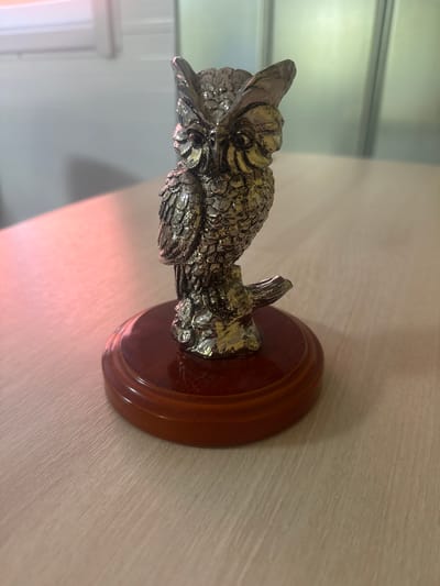 A highly detailed, metallic owl figurine stands on a polished wooden base. The owl is intricately designed, with textured feathers and large, prominent eyes. It is perched on what appears to be a branch. The wooden base is round and has a glossy finish. The figurine is positioned on a light-colored wooden table. In the background, there is a wall with soft lighting, suggesting an indoor environment. The scene is brightly lit, with some light reflecting off the metallic surface of the owl and the wooden base. There are no people or animals present in the image. This image was taken in Sof'ino, Russia.