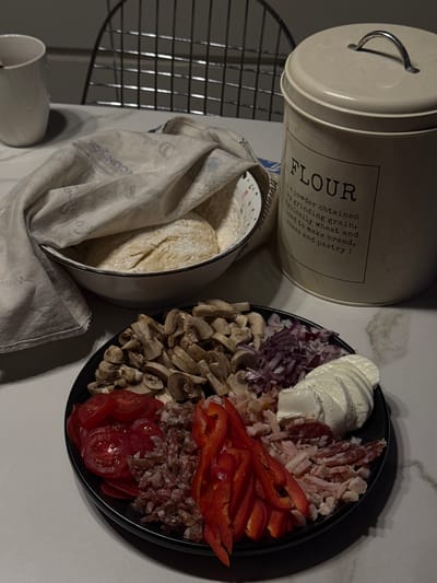 This media file captures a detailed still life of food preparation, likely for baking, set on a kitchen countertop in Ptichnoye, Russia. In the upper left, a cream-colored cylindrical tin with a handle on its lid is prominent. The tin features text including "ΚΟΝΝΠΟΡ" and "FLOUR" in large letters, with a description below: "a powder obtained by grinding grain, typically wheat and used to make bread, cakes and pastry".
To the right, a black circular plate is meticulously arranged with various pizza toppings. These include slices of fresh mozzarella, different types of cured meats such as sliced salami and shredded chicken or ham, vibrant red bell pepper strips, sliced cherry tomatoes, sliced mushrooms, and finely diced red onion.
In the lower-left, a white bowl holds prepared dough, partially covered by a light-colored, possibly flour-dusted, fabric cloth or towel. A metal cooling rack stands vertically behind the bowl, and a white ceramic mug is partially visible below the rack. The lighting is somewhat dim, casting subtle shadows, suggesting an indoor setting, possibly during the evening. The overall scene conveys a sense of organized culinary activity.