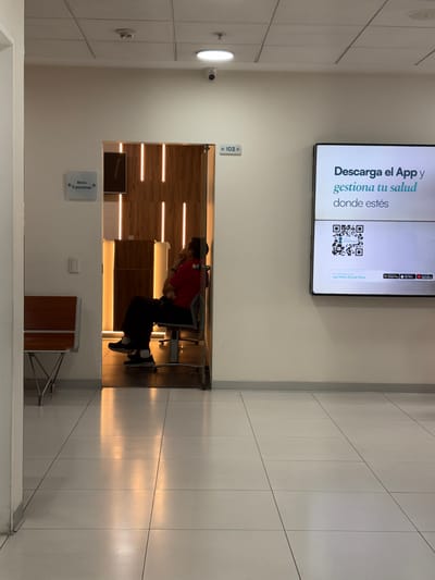 The image shows an indoor scene in what appears to be a medical facility in La Victoria, Peru, based on the Spanish text on the digital display advertising an app for Clínica Ricardo Palma. The interior features tiled flooring and walls. There's a digital display mounted on the wall with a QR code and text promoting an app. Below it, there is a doorway.
Inside the doorway, a person is seated on a metal chair, dressed in a red shirt and black pants. The person is holding their face, possibly in distress. Behind the person, the wall is decorated with horizontal light strips. To the left of the doorway, a sign reads "Aforo 5 personas" (Capacity 5 people). To the right of the doorway, there's a small, wooden stool with metal legs. The environment is brightly lit, suggesting a modern and functional space. A surveillance camera is attached to the ceiling.