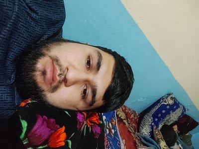A bearded man with dark hair rests on a floral-patterned pillow against a blue and white wall. His eyes are open, looking towards the camera. He wears a dark, patterned shirt. Various blankets and pillows with different patterns and colors are visible in the background.
