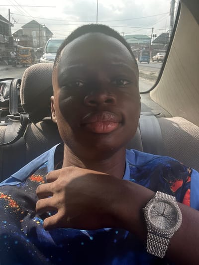 The image displays a young male seated in the rear passenger compartment of a vehicle, facing the camera for a self-portrait. The scene is set in the city of Elimgbu, Nigeria. The subject has dark skin, a short, dark haircut, and a neutral facial expression, looking directly at the viewer. He wears a blue short-sleeved shirt with a vibrant, multi-colored abstract or cosmic pattern. On his left wrist, he wears a large, silver-toned wristwatch heavily encrusted with clear, sparkling stones, featuring Roman numerals on its dial. His left hand is positioned across his chest, partially in shadow.

Behind the subject, a beige fabric headrest is visible. To his left, a black bag with metallic hardware rests on an adjacent seat. Through the vehicle's rear window, an urban daylight street scene is visible under a hazy sky. The background includes multi-story buildings, extensively crisscrossed by overhead utility wires. Traffic includes a white van positioned centrally, a yellow tricycle (keke Napep) on the left, and other indistinct vehicles further down the road. Several indistinct human figures are also visible in the distant background.