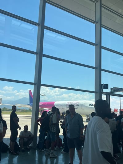 This is a candid, eye-level, vertical shot of a Wizz Air airplane at an airport, likely Tirana International Airport Nënë Tereza, given its proximity to Kruja, Albania. The airplane is pink and white and is parked on the tarmac. In the foreground, a group of people are standing and sitting, presumably waiting to board the flight. The airport terminal has large windows that overlook the tarmac and the airplane. Outside the windows, there are mountains in the distance. The scene is brightly lit by natural daylight. The people in the foreground are casually dressed, and some are looking towards the airplane. The overall atmosphere is one of travel and anticipation.