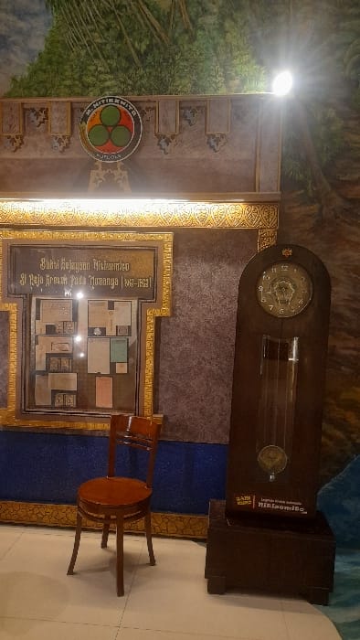 The image displays an indoor exhibit area, identified as being in Glantengan, Indonesia. The scene is dominated by a wall featuring a partial mural of green foliage at the top, transitioning to a textured brown wall below, adorned with gold decorative borders and a lower blue textured section.
Centered at the top of the wall is a circular emblem with "M. NITIKMITO" at its apex and "KUDLOLO" at its base, framing three interconnected green circles on a red background. Below this, on the left side of the frame, is a large, rectangular display case with an ornate gold-colored frame. Inside the case, various historical documents, maps, and papers are arranged and visible. Text above these items reads: "BUKTI KEJAYAAN NITIEMTO" and "DI ERA KEEMASAN PADA MASANYA | 1937-1951".
In front of the display case, a single, light-brown wooden chair with a rounded seat and slatted backrest is positioned on the light-colored tiled floor. To the right of the display case, a tall, dark wooden grandfather clock stands on a darker, block-like base. The clock features a visible pendulum and a clock face with numerals. A small white sign affixed to the clock's base includes text such as "Jati Mulia," "Legenda Kretek Bojongso," and "nitismito.com," indicating a connection to the history of kretek (clove cigarettes). The scene is illuminated by bright overhead spotlights, with one particularly strong light source visible in the upper right quadrant. No individuals are present in the image.