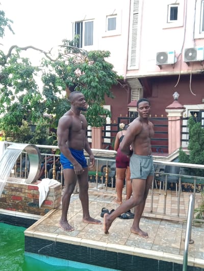 Two shirtless men wearing swim trunks stand on a tiled deck next to a green pool. One man is in blue trunks, while the other is in gray. In the background, a woman in shorts is partially visible. Behind the group is a three-story building with air conditioning units on the exterior. A mature tree provides shade. The setting suggests a private residence or resort with outdoor amenities.