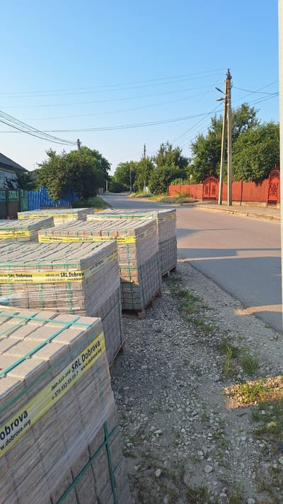 In the city of Sărata-Galbenă, Moldova, on a clear, sunny day, stacks of paving stones are neatly arranged on pallets along a gravelly roadside. The paving stones are wrapped in clear plastic with yellow bands indicating "SRL Dobrova," a company name, and contact information. A paved road runs alongside the paving stones, leading into the distance. On the left side of the road, a residential building with a blue fence is visible, while on the right, a red fence with ornate gates lines the street. Trees and utility poles are scattered throughout the landscape, suggesting a suburban or semi-rural setting. The sky is a bright, cloudless blue, and the sun casts long shadows, indicating it is either early morning or late afternoon.