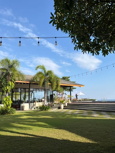 A resort restaurant with a lawn and ocean view is shown in Pasir Putih, Indonesia. The restaurant has large windows and is decorated with string lights. There are palm trees and a playground on the property. In the foreground, a lush green lawn is illuminated by sunlight, casting shadows across the grass. A woman in a dark dress is standing near a white umbrella, while another person in light clothing stands nearby. The ocean is visible in the background, beyond a tiled patio area with seating.