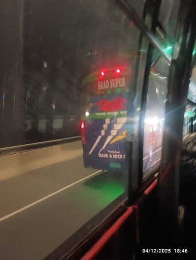 The image depicts the rear exterior of a blue passenger bus, labeled "SAAD SUPER," viewed from inside another vehicle through a window, at approximately 18:46 on December 4, 2025, in Naodoba, Bangladesh. The scene is illuminated by artificial light, suggesting nighttime or late dusk.

The "SAAD SUPER" bus occupies the central and right portions of the frame. Its rear is predominantly blue with a large white and yellow lightning bolt graphic on the lower section. Above this, a red, abstract bird-like logo is visible. Red text spelling "SAAD SUPER" is present near the top, illuminated by internal or external lights. Below the lightning bolt, partially obscured, are the words "HAVE A NICE T" in white lettering. Two red taillights are active on the bus. A distinct green light is emitted or reflected onto the asphalt road surface directly beneath the bus's rear bumper.

The viewing vehicle's interior is visible in the foreground, showing a red horizontal bar, possibly a handrail or part of a seat structure, along the bottom edge of the frame. Vertical dark structural elements of the window frame are visible on the right. The window itself shows reflections and minor smudges.

The road is an asphalt surface with a clear white lane marking visible on the left side, indicating multiple lanes. A concrete barrier runs along the left edge of the road, separating it from the dark background. The environment is dark, with distant artificial lights visible on the far right. No individuals are discernibly present within the visible area. Both vehicles appear to be in motion.