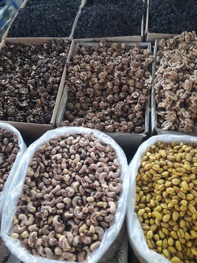 The image presents a close-up, overhead view of a market stall displaying various bulk nuts and dried fruits in Ghafurov, Tajikistan. The scene is dominated by multiple types of these goods, arranged in different containers.

On the left side, there are several rectangular cardboard boxes filled with dark, dried produce, possibly black raisins or dried mulberries. To their right, more cardboard boxes hold abundant quantities of shelled walnuts, showcasing their intricate, natural patterns.

The right side of the frame features large clear plastic bags, each filled with a different type of nut. The uppermost bag on the right is packed with light green, mostly shelled pistachios. Below this, another large plastic bag is brimming with cashews, which appear to be either raw or lightly roasted, characterized by their varied light brown and earthy tones. A portion of a third plastic bag, also containing cashews, is visible at the bottom right.

The arrangement of items in distinct containers suggests a commercial setting, likely a vibrant market or a shop, where these goods are sold. There are no people visible in the frame, and no discernible text can be seen on any of the packaging or labels. The natural appearance of the nuts and dried fruits implies a focus on fresh or unprocessed quality.