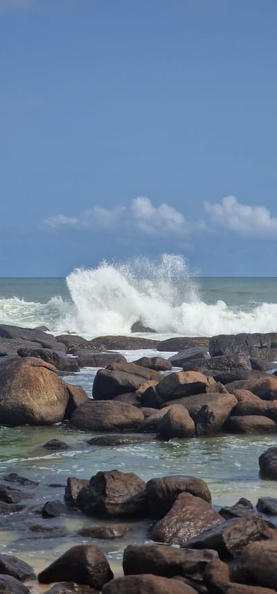 The image shows a rocky shoreline in Dondra, Sri Lanka. Large, dark rocks are clustered along the water's edge. A wave is crashing against the rocks, creating white spray. The sky is blue with scattered white clouds. The water is a mix of green and blue.