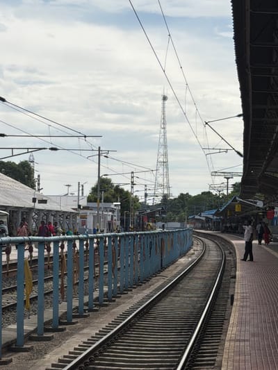 The image depicts a busy train station platform in Thanjavur, India. Several people are visible on the platform, some standing and others appearing to be walking. The scene is set outdoors under a cloudy sky, suggesting it is daytime.
Dominant objects in the scene include train tracks, overhead electrical wires, and a tall metal tower in the distance, likely a communication tower. A blue metal railing with wooden planks runs alongside the tracks, separating the platform from the tracks themselves. Various structures that appear to be part of the station's infrastructure, such as pillars and sheds, are also present. The platform surface is paved with reddish-brown bricks.
The composition of the image, with its emphasis on the curved train tracks and the overhead wires, creates a sense of depth and activity. The presence of people suggests a normal day at the station, with commuters or travelers possibly waiting for trains or disembarking. The lighting indicates it is likely midday or afternoon, with diffused sunlight due to the clouds. There is no visible text in the image.