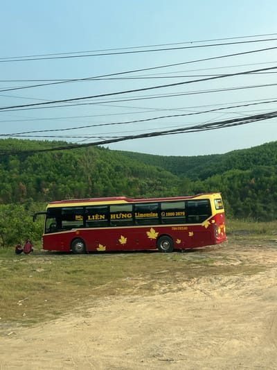 A red and yellow double-decker bus, adorned with maple leaf decals, is parked on a dirt and grassy shoulder next to a road. The bus has the words "LIEN HUNG Limousine" and a phone number (1900 2679) printed on its side. The license plate number is 79H-093.90. In the background, there is a lush green hillside and a clear blue sky. Power lines run diagonally across the left side of the frame. Two people wearing dark clothing are visible near the front of the bus. The location is in Vietnam.