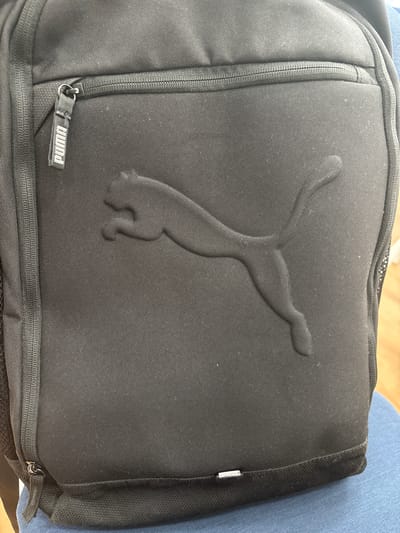 The image shows a close-up of a black Puma backpack. The backpack has a prominent embossed Puma logo on the front. A zipper with a Puma branded pull tab is visible at the top of the backpack. The backpack is resting on a blue surface, possibly a chair. The location is not discernible from the image.