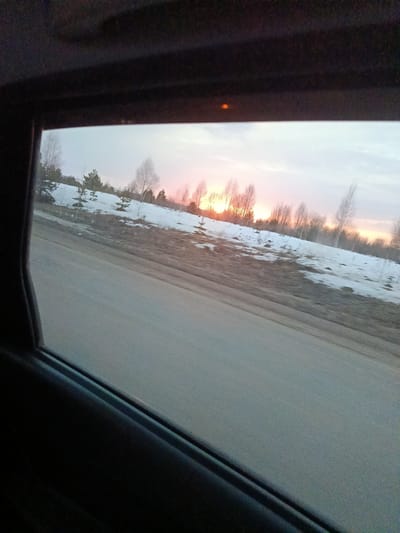 The photograph, presumably taken from inside a moving vehicle, shows a landscape scene. The view is framed by the vehicle's window and interior trim. Outside, a road stretches across the lower part of the frame. Beyond the road, a snow-covered hillside with sparse vegetation and trees is visible. The sky shows a sunset or sunrise with an orange glow near the horizon.
There is no definitive evidence to confirm the location is specifically Koliushevo, Russia, although it is a plausible setting based on the landscape. There are no people visible in the image. The primary subjects are the road, the snow-covered landscape, and the sky at sunset/sunrise. No interactions are observable.